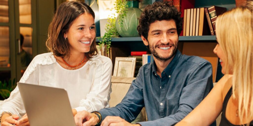 Three colleagues smile and chat around a laptop in a cozy workspace with shelves and plants in the background.